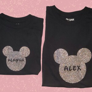 May include: Two black t-shirts with a silver glitter Mickey Mouse head design. One shirt says "ALAYNA" and the other says "ALEX".