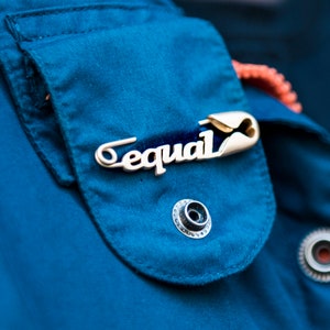 Op de afbeelding: Een close-up van een blauw jack met een zilveren pin die "gelijk" zegt.