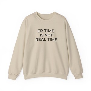 以下が含まれることがあります： ベージュのクルーネックのスウェットシャツ。黒い文字で「ER TIME IS NOT REAL TIME」とプリントされています。長袖トップスは柔らかい素材でできており、カジュアルウェアに適しています。