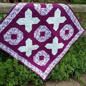 May include: A purple and white quilt with a geometric pattern. The quilt is made with a floral fabric and has a white border.