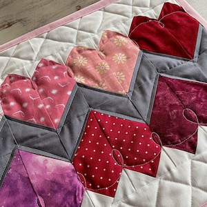 Puede incluir: Un camino de mesa acolchado blanco con un borde rosa presenta un patrón de corazones en varios tonos de rosa, rojo y morado. Los corazones están hechos de diferentes telas con varios patrones, incluyendo lunares, estampados florales y un diseño similar a la acuarela.