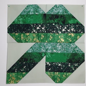 May include: A green and gold patchwork quilt block with a geometric design. The block is made up of several different fabrics, including a green fabric with a white speck pattern, a solid green fabric, and a gold fabric with a green speck pattern.
