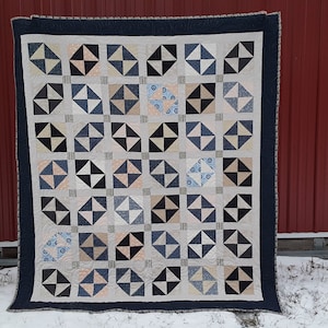 May include: A quilt with a geometric pattern in shades of blue, black, white, and beige. The quilt is made up of squares with a diamond shape in the center. The quilt is hanging on a red barn.