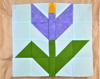 May include: A quilt block featuring a purple tulip with a yellow center and green leaves on a light blue background.