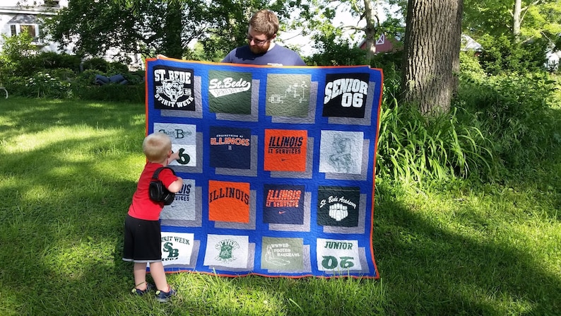 May include: A handmade quilt made from various t-shirts, featuring a blue border and a grid pattern. The t-shirts display text and logos, including "ST. BEDE", "ILLINOIS", and "SENIOR 06". The quilt is held up by two people outdoors.