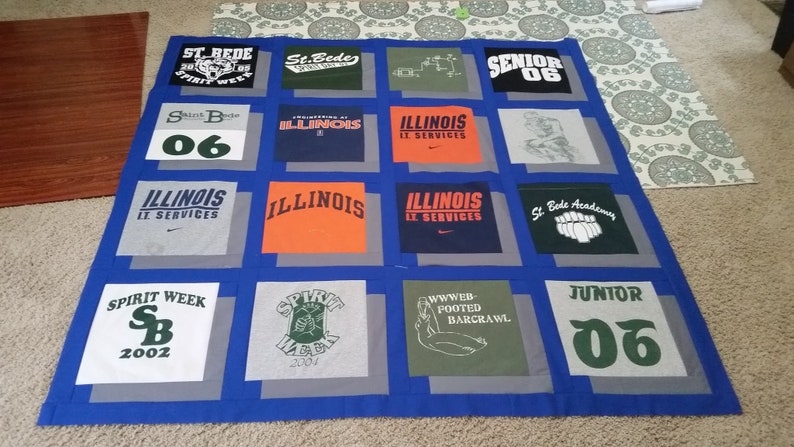 May include: A handmade quilt featuring squares of various t-shirts. The shirts display text and graphics, including "ILLINOIS," "SPIRIT WEEK," and "SENIOR 06." The quilt has a blue border and gray squares, showcasing a unique, personalized design.