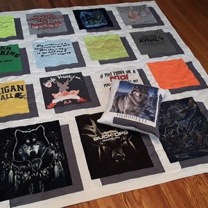 May include: A handmade quilt made from various t-shirts, featuring a variety of graphics and text. The quilt is white with gray borders around each t-shirt square. A decorative pillow with a wolf image rests on the quilt.