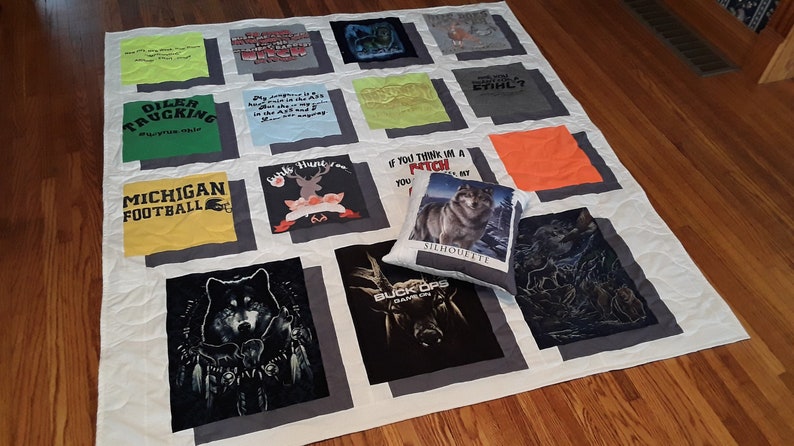 May include: A handmade quilt made from various t-shirts, featuring a variety of graphics and text. The quilt is white with gray borders around each t-shirt square. A decorative pillow with a wolf image rests on the quilt.