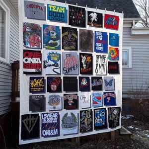 May include: A handmade quilt made from various graphic t-shirts. The shirts feature band names, logos, and text like "ONEONTA STATE 89", "OF MICE & MEN", and "Wiz Khalifa". The quilt is displayed outdoors, showcasing a unique collection.