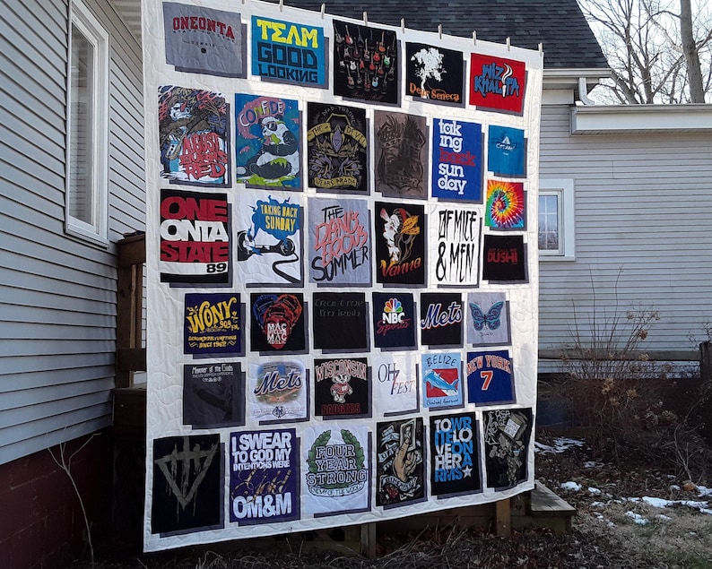 May include: A handmade quilt made from various graphic t-shirts. The shirts feature band names, logos, and text like "ONEONTA STATE 89", "OF MICE & MEN", and "Wiz Khalifa". The quilt is displayed outdoors, showcasing a unique collection.
