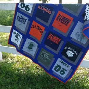 May include: A handmade quilt featuring squares of various t-shirts, primarily in gray, orange, and blue. The quilt has a blue border with a red trim and is draped over a white fence. The t-shirts display text and logos.