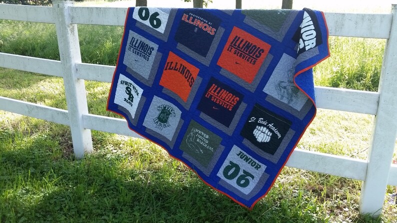 May include: A handmade quilt featuring squares of various t-shirts, primarily in gray, orange, and blue. The quilt has a blue border with a red trim and is draped over a white fence. The t-shirts display text and logos.