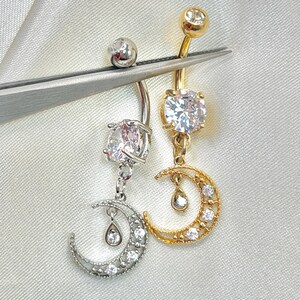 May include: Two belly button rings, one silver-toned and one gold-toned, each featuring a round clear gemstone at the top and a crescent moon charm dangling below. The moon charms are accented with small clear stones and a tiny teardrop-shaped charm.