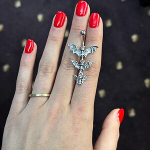 May include: A silver body piercing with three bat charms hanging from it. The piercing is held in a hand with red nail polish.