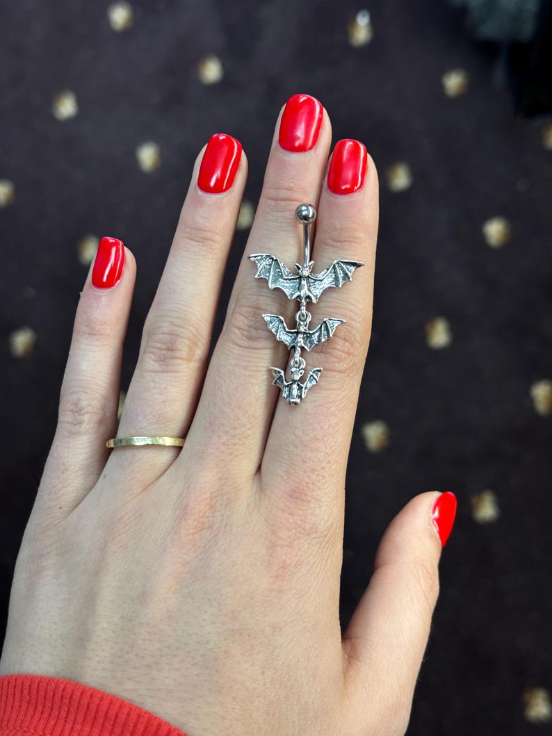 May include: A silver body piercing with three bat charms hanging from it. The piercing is held in a hand with red nail polish.