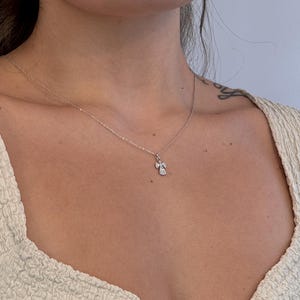 May include: A delicate silver necklace with a small, sparkling angel pendant. The pendant features a detailed design with clear gemstones, suspended from a fine chain. The necklace is worn against a light-colored top.