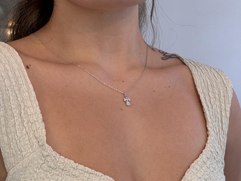 May include: A delicate silver necklace with a small, sparkling angel pendant. The pendant features a detailed design with clear gemstones, suspended from a fine chain. The necklace is worn against a light-colored top.