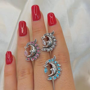 Puede incluir: Tres anillos de tono plateado con un diseño de cara de luna creciente. Cada anillo está adornado con pequeñas piedras brillantes a lo largo del borde de la luna y una gema colgante de corte marquesa. Un anillo tiene piedras rosas, otro piedras claras y otro piedras azules.