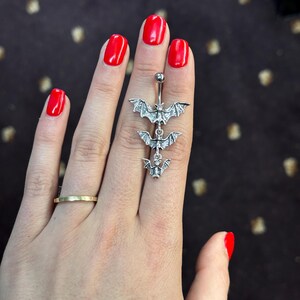 May include: A silver body piercing with three dangling bat charms. The bats are detailed with wings and have a shiny finish.