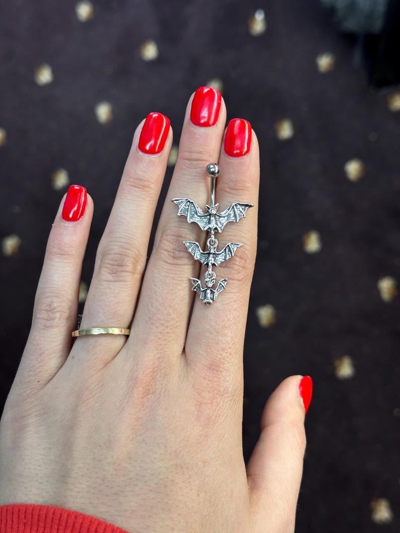 May include: A silver body piercing with three dangling bat charms. The bats are detailed with wings and have a shiny finish.