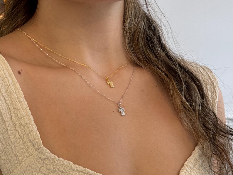 May include: Two delicate necklaces with angel pendants. One necklace is gold, the other is silver. The angel pendants are small and detailed, with a sparkling appearance. The necklaces are layered, creating a stylish and elegant look.