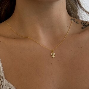 May include: A delicate gold necklace featuring a small angel pendant. The pendant is adorned with sparkling gemstones, adding a touch of elegance. The necklace is displayed against a skin tone background, highlighting the jewelry's design.