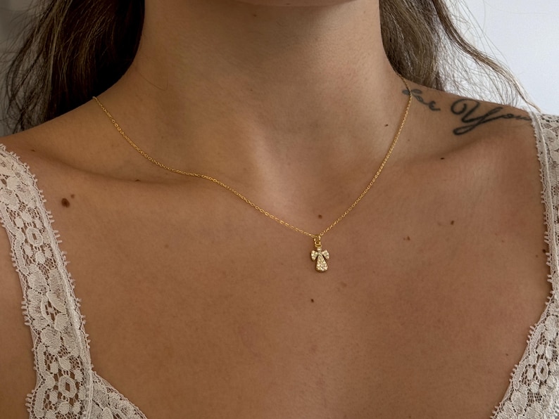 May include: A delicate gold necklace featuring a small angel pendant. The pendant is adorned with sparkling gemstones, adding a touch of elegance. The necklace is displayed against a skin tone background, highlighting the jewelry's design.