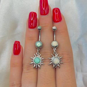 May include: Two silver-toned navel rings featuring a curved barbell design. Each piece has a small round stone at the top and a larger sun-shaped charm at the bottom. One sun charm contains a light blue stone, while the other contains a white stone.