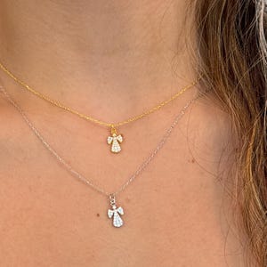 May include: Two delicate necklaces with angel pendants. The top necklace is gold, and the bottom is silver. Each pendant features a small angel design with sparkling details, creating a layered look. The necklaces are worn close to the neck.