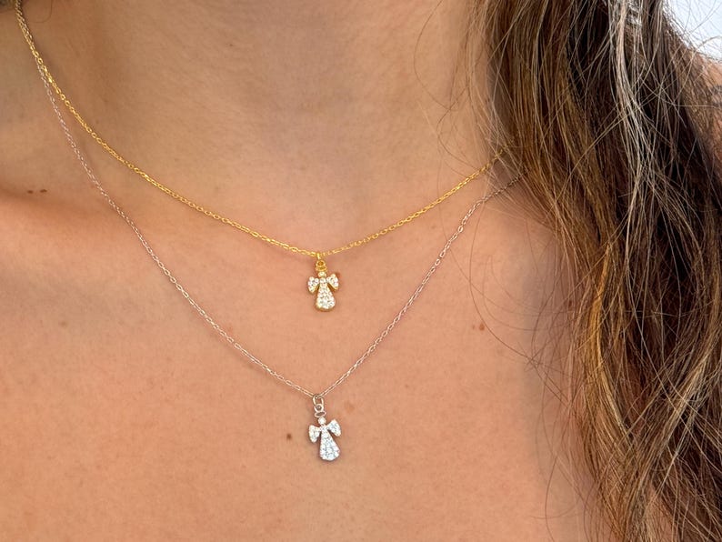 May include: Two delicate necklaces with angel pendants. The top necklace is gold, and the bottom is silver. Each pendant features a small angel design with sparkling details, creating a layered look. The necklaces are worn close to the neck.