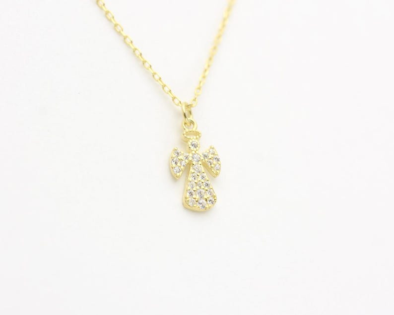 May include: A gold-colored angel pendant necklace. The angel pendant is adorned with small, clear gemstones. The necklace has a delicate chain and is set against a white background. The jewelry is suitable for everyday wear.
