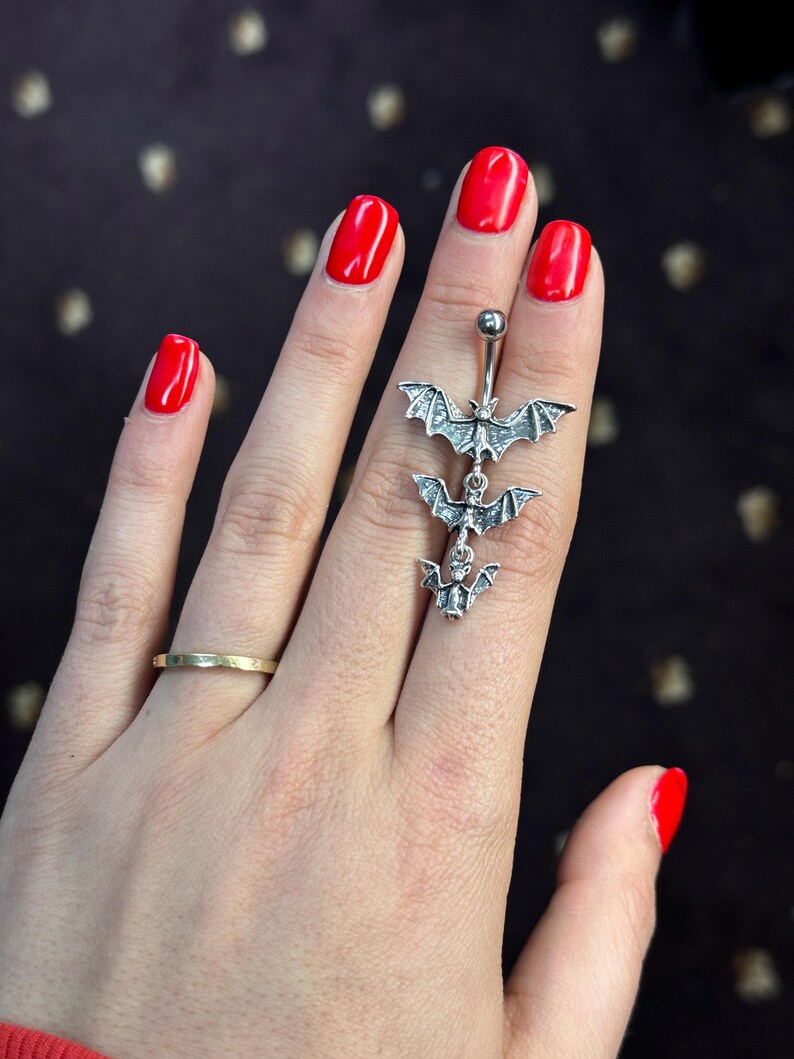May include: A silver belly button ring with three dangling bat charms. The ring is on a person's finger with red nail polish.