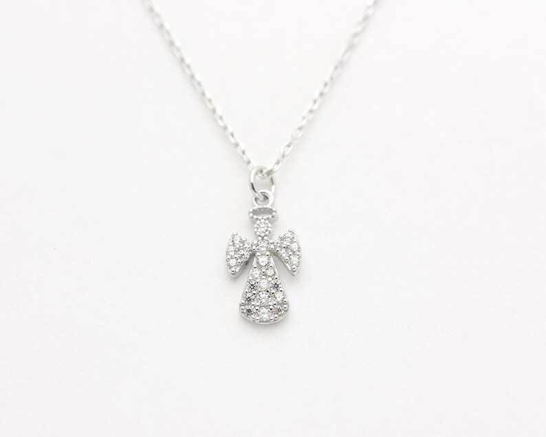 May include: A silver-colored angel pendant necklace. The angel pendant is encrusted with small, clear gemstones. The necklace chain is delicate and silver. The background is plain white.
