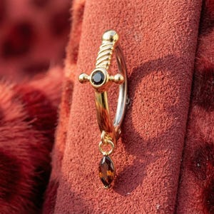 Może przedstawiać: Złoty i srebrny kolczyk z motywem miecza. Kolczyk posiada czarny kamień szlachetny i wiszący brązowy kamień szlachetny. Kolczyk jest osadzony na czerwonym tle z nadrukiem w cętki.