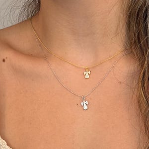 May include: Two delicate necklaces with angel pendants. One necklace is gold, the other is silver. Each pendant features a small angel design with sparkling details. The necklaces are layered, creating a stylish and elegant look.