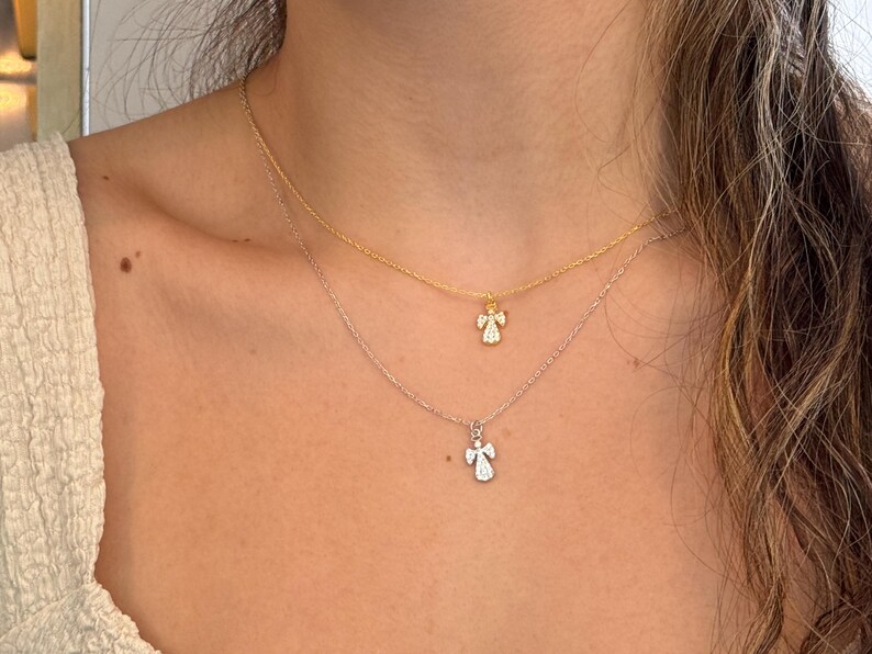 May include: Two delicate necklaces with angel pendants. One necklace is gold, the other is silver. Each pendant features a small angel design with sparkling details. The necklaces are layered, creating a stylish and elegant look.