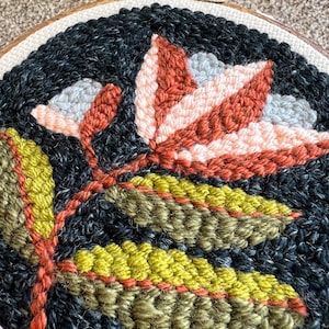 K&ouml;nnte beinhalten: Nahaufnahme eines runden Stickrahmens mit einem floralen Design. Das Design zeigt eine gro&szlig;e, stilisierte Blume mit orangefarbenen, rosa und grauen Bl&uuml;tenbl&auml;ttern. Die Blume ist von gr&uuml;nen Bl&auml;ttern umgeben.