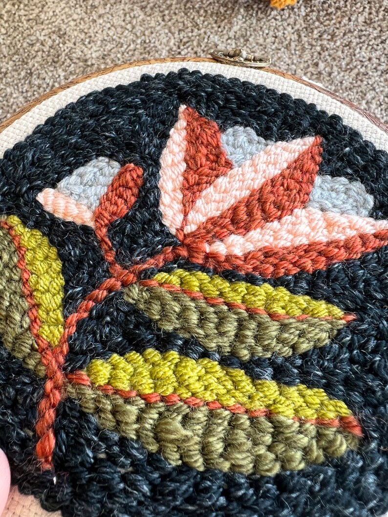 K&ouml;nnte beinhalten: Nahaufnahme eines runden Stickrahmens mit einem floralen Design. Das Design zeigt eine gro&szlig;e, stilisierte Blume mit orangefarbenen, rosa und grauen Bl&uuml;tenbl&auml;ttern. Die Blume ist von gr&uuml;nen Bl&auml;ttern umgeben.