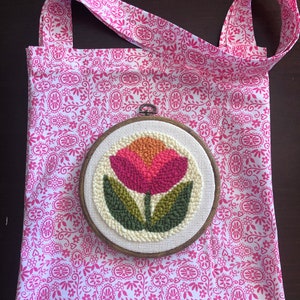 May include: A white tote bag with a pink floral pattern and a wooden embroidery hoop with a pink and green flower design.