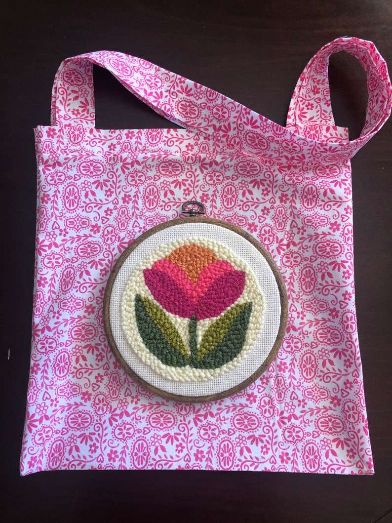 May include: A white tote bag with a pink floral pattern and a wooden embroidery hoop with a pink and green flower design.
