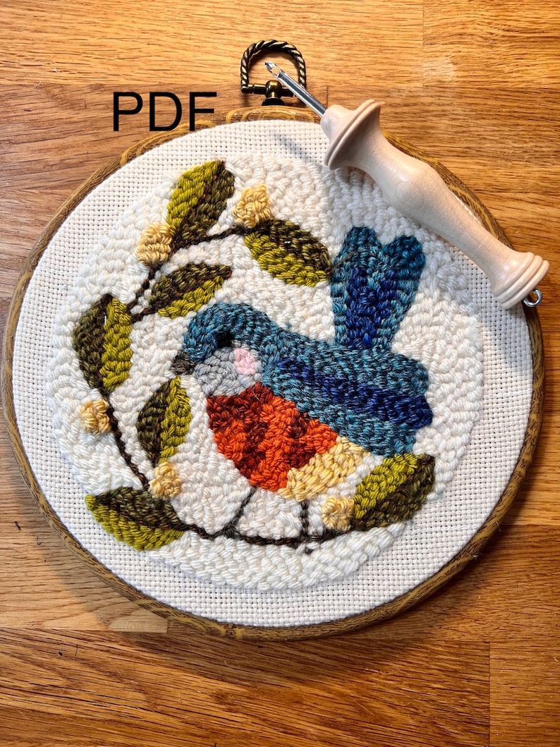 May include: A completed punch needle embroidery of a blue and orange bird on a branch with green leaves and yellow flowers. The embroidery is in a wooden hoop with a metal hanger. A punch needle tool rests on the hoop. The word "PDF" is in the upper left corner.