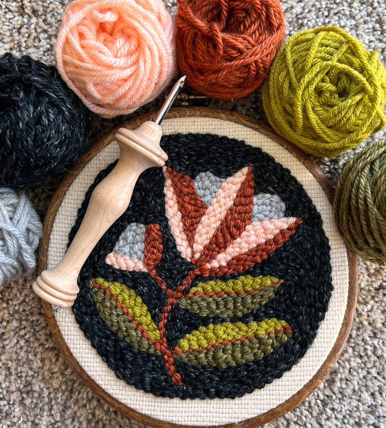 K&ouml;nnte beinhalten: Nahaufnahme eines fertigen Nadelbildes mit einer stilisierten Blume mit Bl&auml;ttern in Gr&uuml;n-, Braun-, Rosa- und Graut&ouml;nen. Das Nadelbild ist in einem Holzrahmen gespannt und von Garnkn&auml;ueln in verschiedenen Farben umgeben.