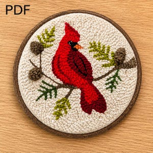 May include: A round, hand-stitched textile art piece featuring a vibrant red cardinal bird perched on a branch with green needles and brown pinecones. The artwork is set against a cream-colored background and framed with a brown border. The text "PDF" is in the upper left corner.