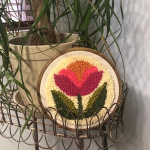 May include: A wooden hoop with a cream-colored canvas featuring a pink, orange, and yellow flower with green leaves. The flower is made of yarn and is a punch needle embroidery.