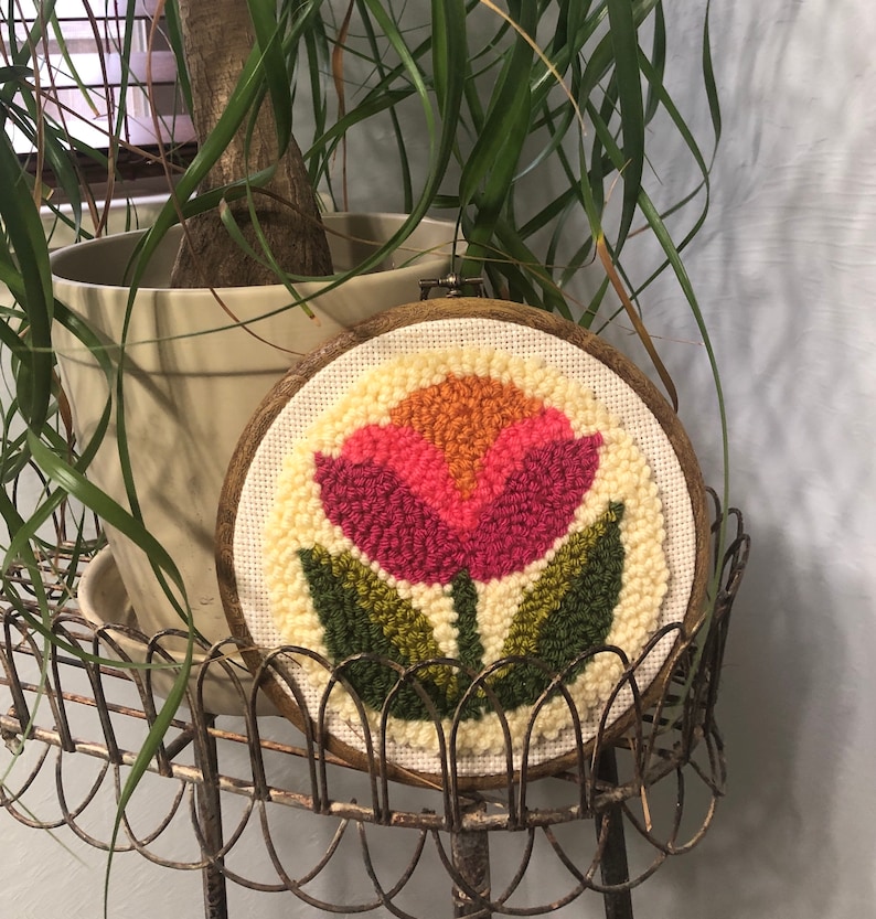 May include: A wooden hoop with a cream-colored canvas featuring a pink, orange, and yellow flower with green leaves. The flower is made of yarn and is a punch needle embroidery.