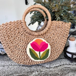May include: A straw tote bag with a round, white patch featuring a pink and orange flower with green leaves. The bag has a woven texture and two brown handles.