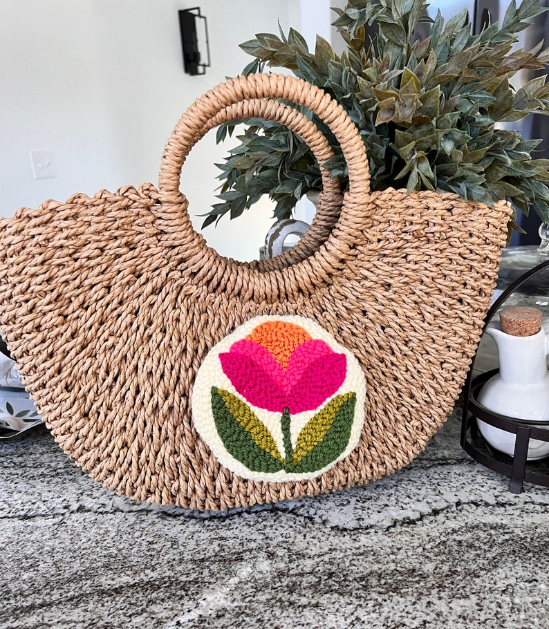 May include: A straw tote bag with a round, white patch featuring a pink and orange flower with green leaves. The bag has a woven texture and two brown handles.
