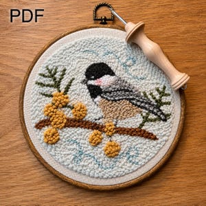Peut inclure: Une œuvre d'art au point de croix circulaire encadrée, représentant un oiseau sur une branche avec des baies jaunes. L'oiseau a des plumes noires, blanches et marron. Un outil de punch needle en bois est posé sur le cadre. Le mot "PDF" est visible.