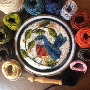 May include: A completed punch needle embroidery of a bluebird with orange breast and blue wings, surrounded by green leaves and white flowers. The embroidery is in a wooden hoop, surrounded by various coloured yarn skeins and a punch needle tool.