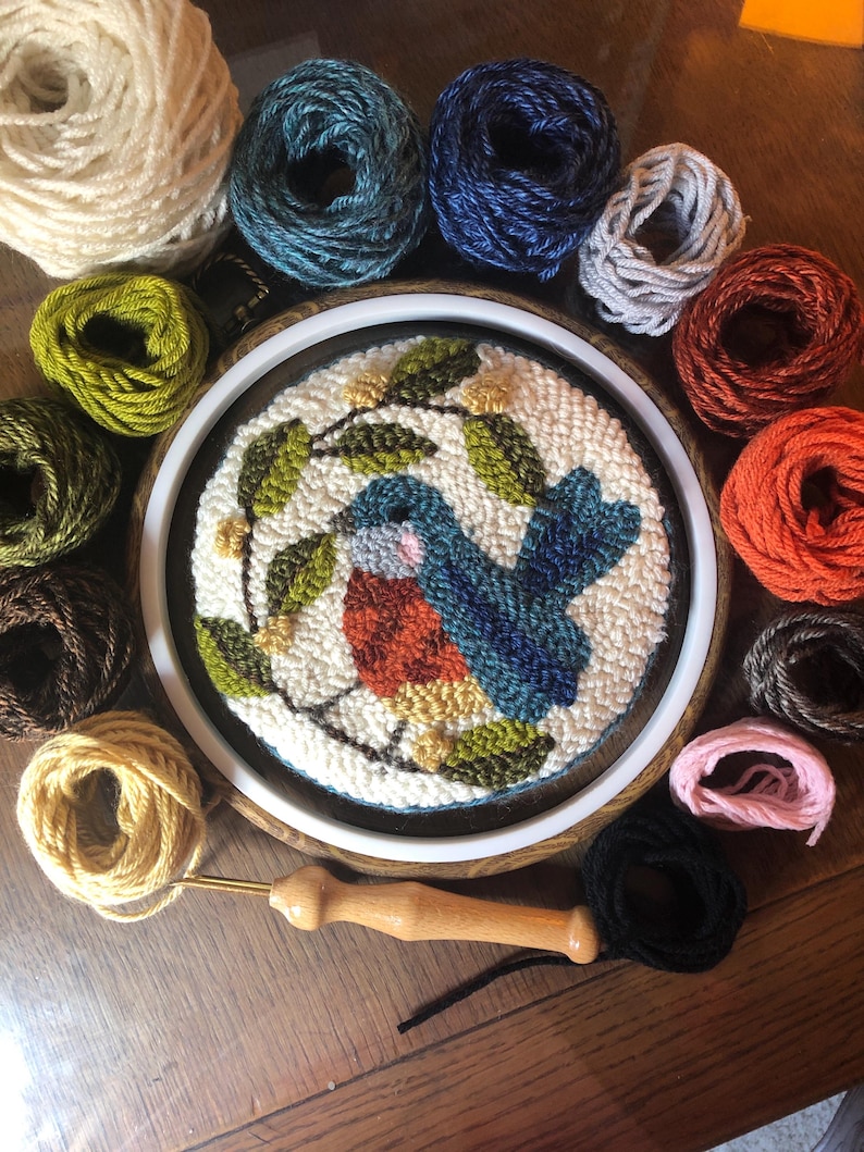 May include: A completed punch needle embroidery of a bluebird with orange breast and blue wings, surrounded by green leaves and white flowers. The embroidery is in a wooden hoop, surrounded by various coloured yarn skeins and a punch needle tool.